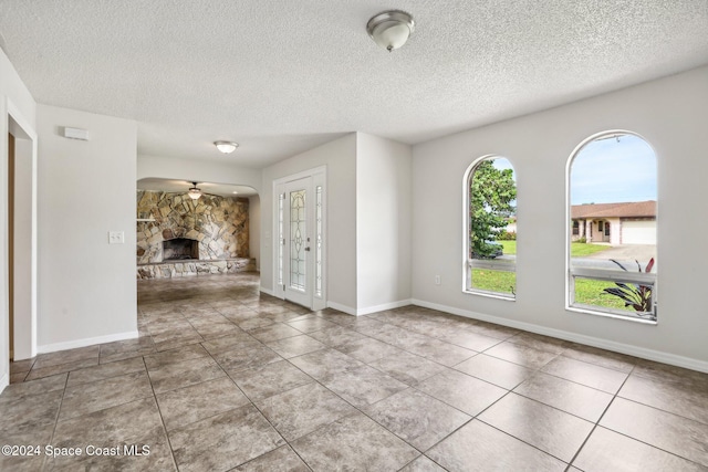 unfurnished living room featuring a ceiling fan, a textured ceiling, a stone fireplace, tile patterned floors, and baseboards