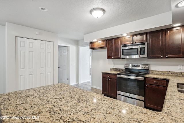 kitchen with stainless steel appliances, light stone counters, tile patterned floors, dark brown cabinetry, and a textured ceiling