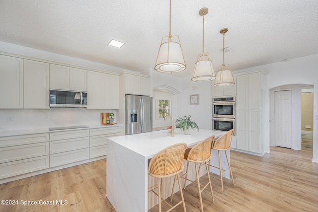 kitchen with stainless steel appliances, sink, light hardwood / wood-style floors, hanging light fixtures, and an island with sink