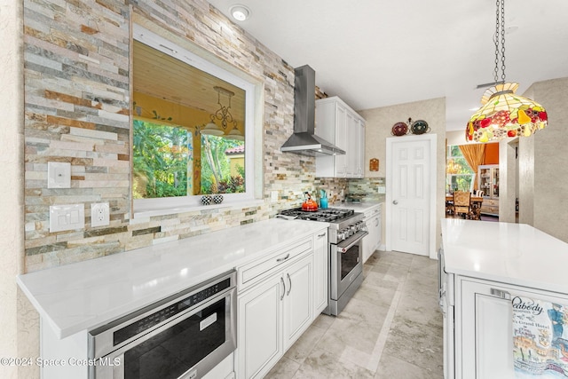 kitchen with wall chimney range hood, tasteful backsplash, white cabinetry, decorative light fixtures, and stainless steel appliances