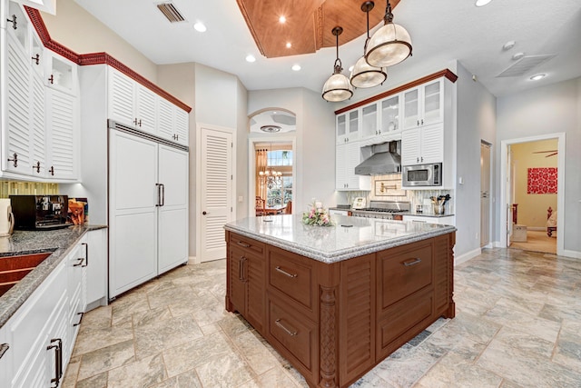 kitchen featuring wall chimney range hood, built in appliances, stone finish floor, white cabinetry, and visible vents