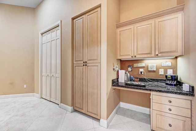 kitchen featuring dark stone counters, baseboards, built in desk, and light brown cabinets
