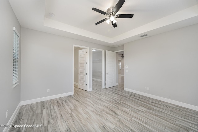 unfurnished bedroom with a closet, light hardwood / wood-style flooring, a tray ceiling, a walk in closet, and ceiling fan