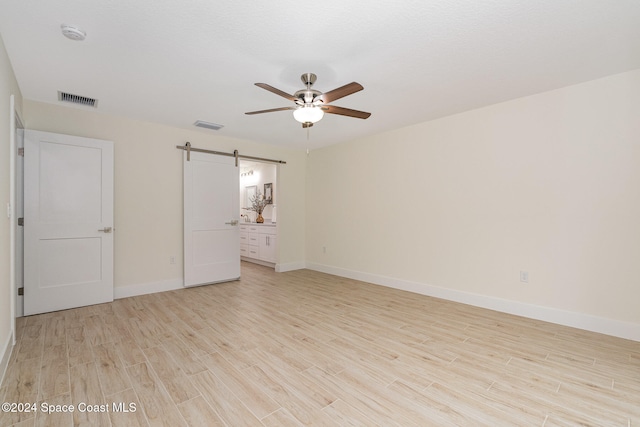 unfurnished bedroom featuring light hardwood / wood-style floors, a barn door, connected bathroom, and ceiling fan