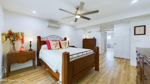 bedroom featuring light hardwood / wood-style floors, a wall unit AC, a barn door, connected bathroom, and ceiling fan