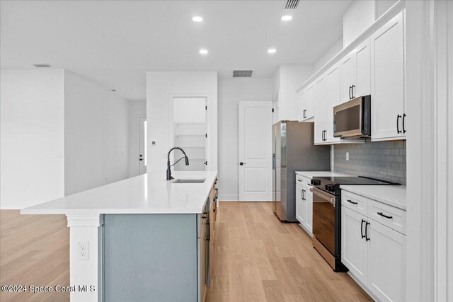 kitchen with appliances with stainless steel finishes, a sink, light wood finished floors, a center island with sink, and backsplash