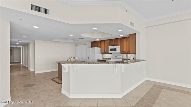 kitchen with a kitchen bar, dark stone counters, kitchen peninsula, ornamental molding, and white appliances