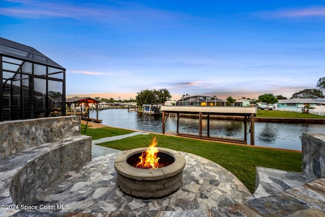 patio terrace at dusk with a lanai, a lawn, a water view, and an outdoor fire pit