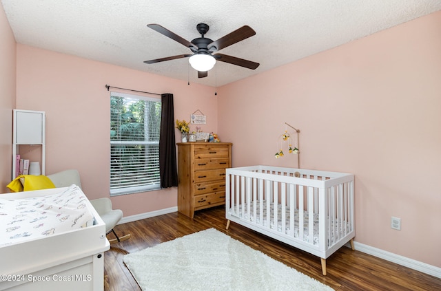 bedroom with a textured ceiling, a nursery area, dark wood-type flooring, and ceiling fan