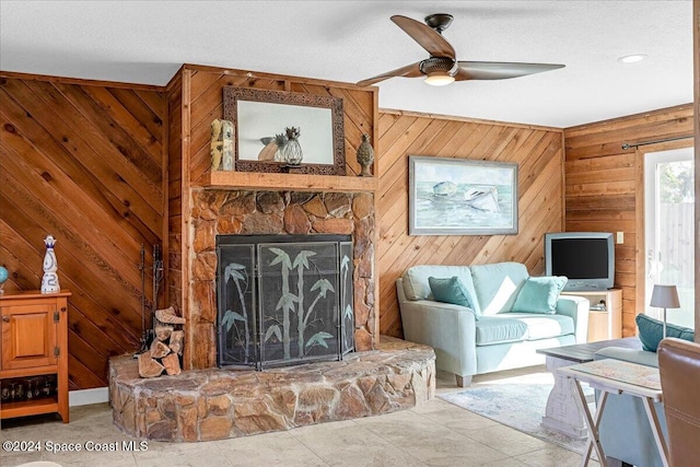 living room featuring a textured ceiling, a stone fireplace, ceiling fan, and wooden walls