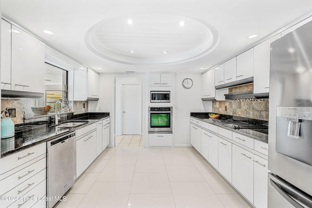 kitchen featuring appliances with stainless steel finishes, a tray ceiling, sink, dark stone countertops, and white cabinetry