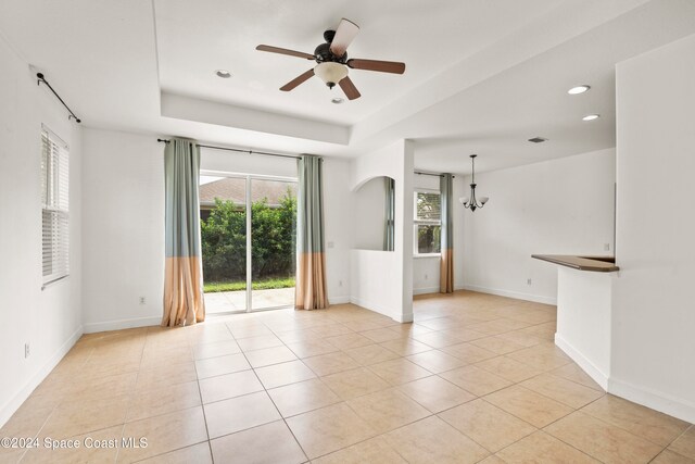 tiled empty room with ceiling fan with notable chandelier and a tray ceiling