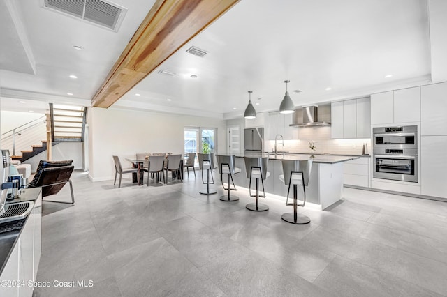 kitchen with pendant lighting, wall chimney range hood, stainless steel double oven, an island with sink, and white cabinetry