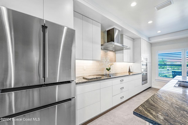 kitchen featuring tasteful backsplash, dark stone counters, stainless steel appliances, wall chimney range hood, and white cabinetry