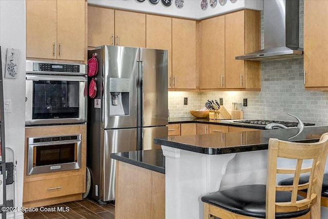 kitchen featuring dark wood-type flooring, a kitchen breakfast bar, wall chimney exhaust hood, appliances with stainless steel finishes, and kitchen peninsula