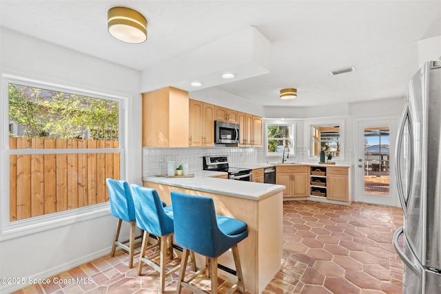 kitchen featuring tasteful backsplash, light brown cabinetry, appliances with stainless steel finishes, a kitchen bar, and kitchen peninsula