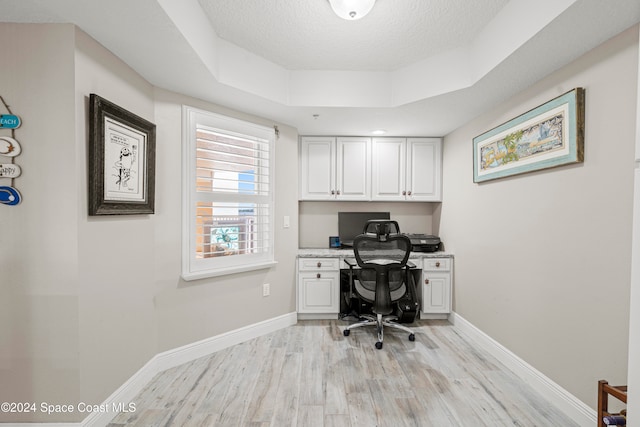 home office with a raised ceiling, light wood-type flooring, built in desk, and a textured ceiling