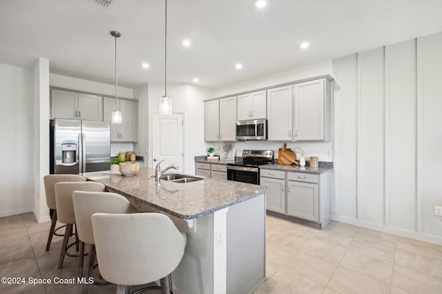 kitchen with light stone counters, stainless steel appliances, sink, decorative light fixtures, and a center island with sink