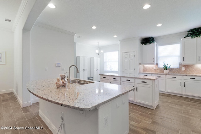 kitchen with light stone counters, a kitchen island with sink, crown molding, sink, and white cabinetry