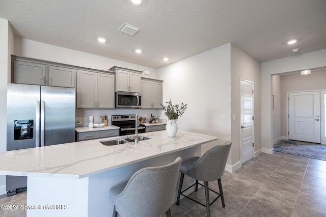 kitchen featuring appliances with stainless steel finishes, light stone counters, a textured ceiling, a center island with sink, and gray cabinets