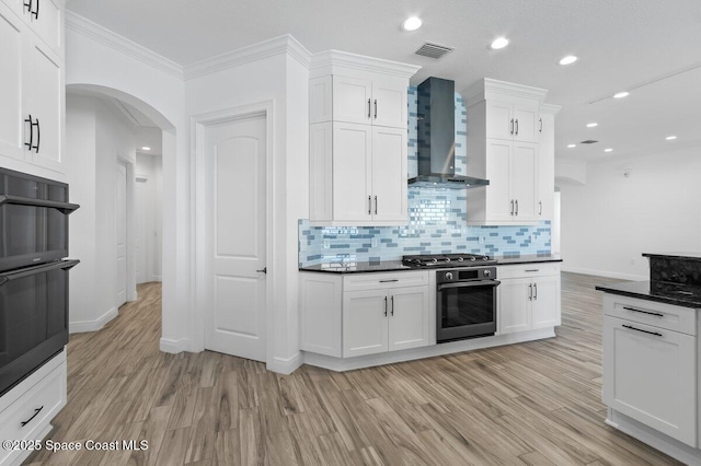 kitchen featuring dark countertops, visible vents, wall chimney range hood, light wood-style floors, and arched walkways