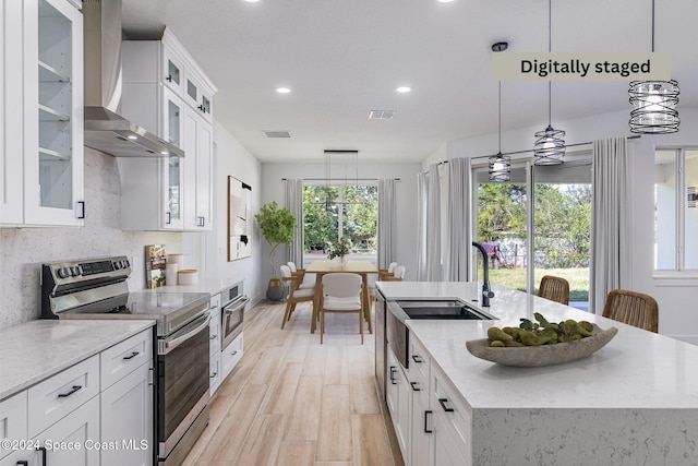 kitchen with appliances with stainless steel finishes, wall chimney exhaust hood, a center island with sink, white cabinetry, and hanging light fixtures