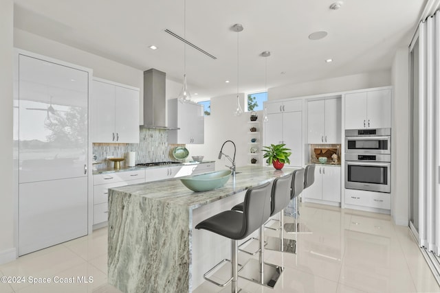 kitchen with white cabinets, a center island with sink, wall chimney range hood, sink, and decorative light fixtures