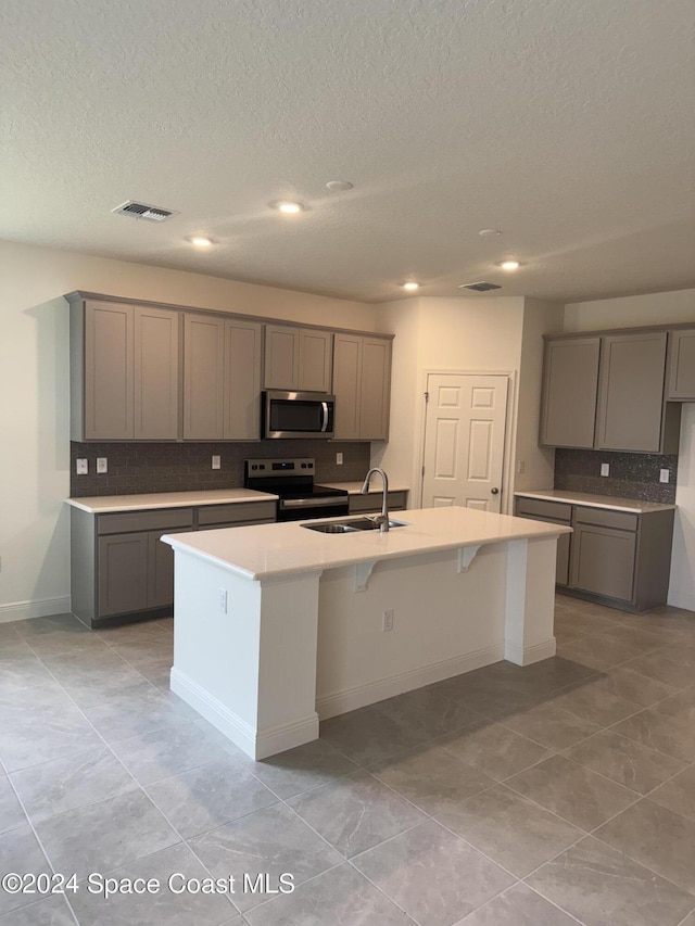 kitchen with gray cabinetry, sink, decorative backsplash, a kitchen island with sink, and appliances with stainless steel finishes