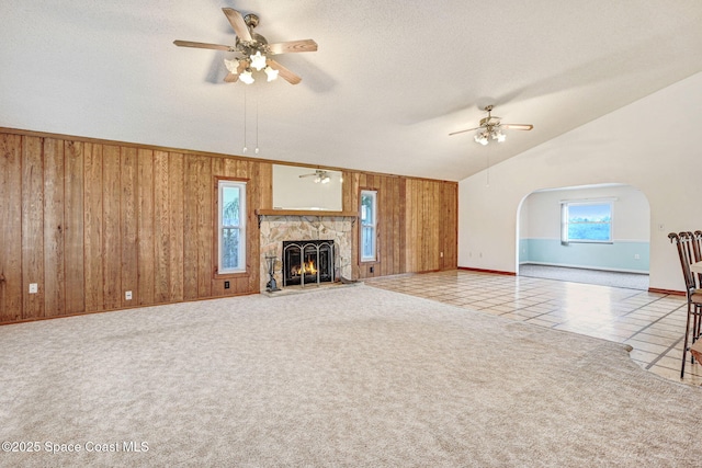 unfurnished living room featuring ceiling fan, vaulted ceiling, tile patterned floors, wooden walls, and a stone fireplace