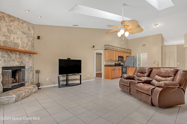 living room with high vaulted ceiling, a stone fireplace, a skylight, ceiling fan, and light tile patterned floors