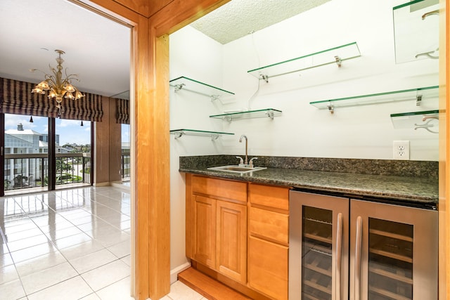 bar with dark stone counters, beverage cooler, sink, light tile patterned floors, and an inviting chandelier