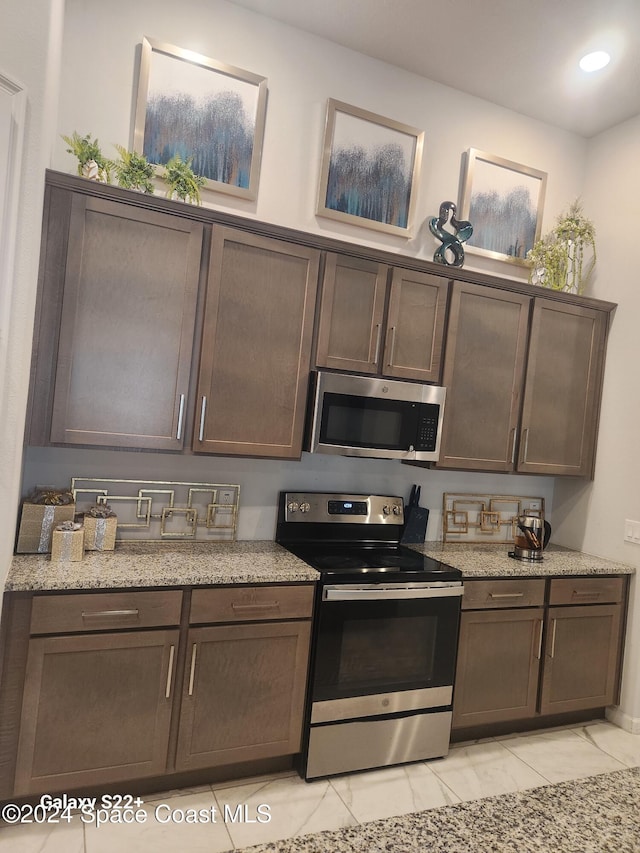 kitchen featuring dark brown cabinetry, light stone counters, light tile patterned floors, and appliances with stainless steel finishes