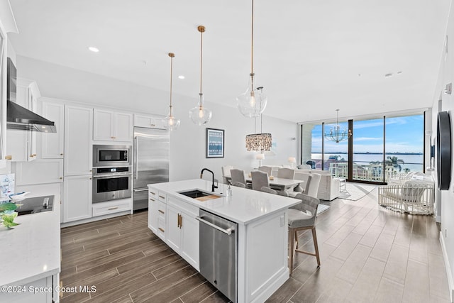 kitchen with pendant lighting, a kitchen island with sink, sink, built in appliances, and white cabinetry