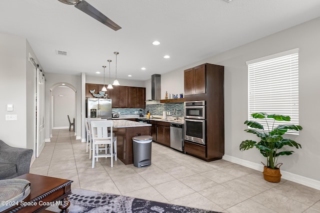 kitchen featuring appliances with stainless steel finishes, wall chimney range hood, a center island, hanging light fixtures, and a breakfast bar area