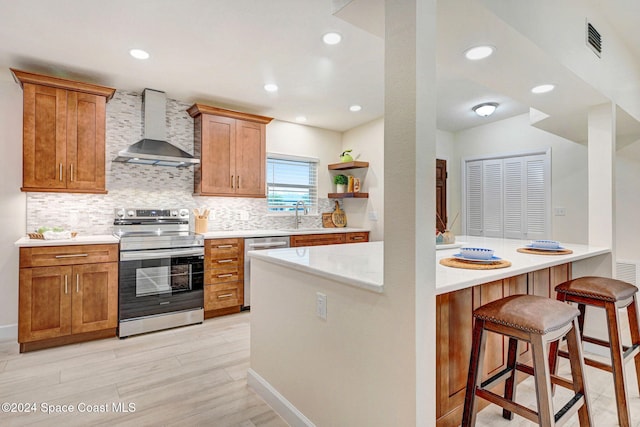 kitchen featuring kitchen peninsula, appliances with stainless steel finishes, light wood-type flooring, wall chimney range hood, and a breakfast bar area