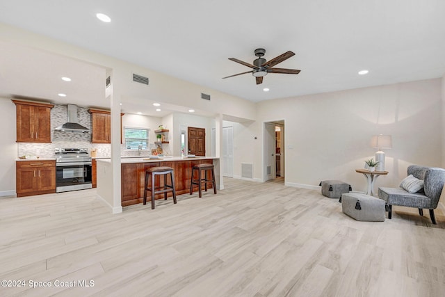 interior space featuring a kitchen bar, wall chimney exhaust hood, light hardwood / wood-style floors, ceiling fan, and stainless steel stove