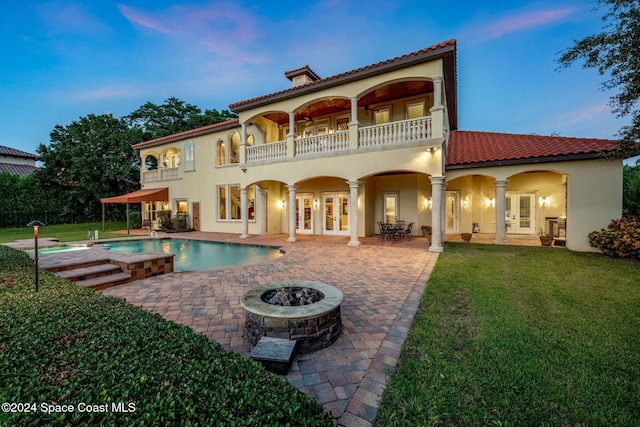 back house at dusk featuring a fire pit, french doors, a swimming pool with hot tub, a balcony, and a patio