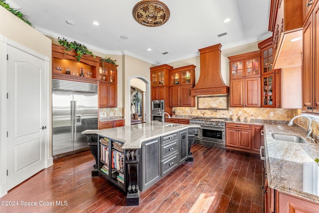 kitchen featuring a kitchen island with sink, sink, built in appliances, light stone countertops, and custom range hood