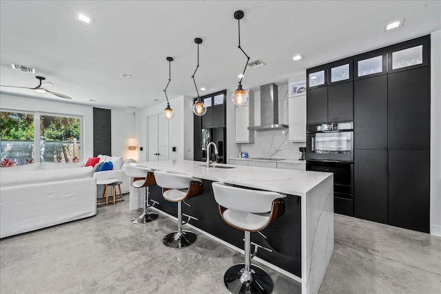 kitchen with wall chimney range hood, a large island with sink, pendant lighting, decorative backsplash, and a breakfast bar