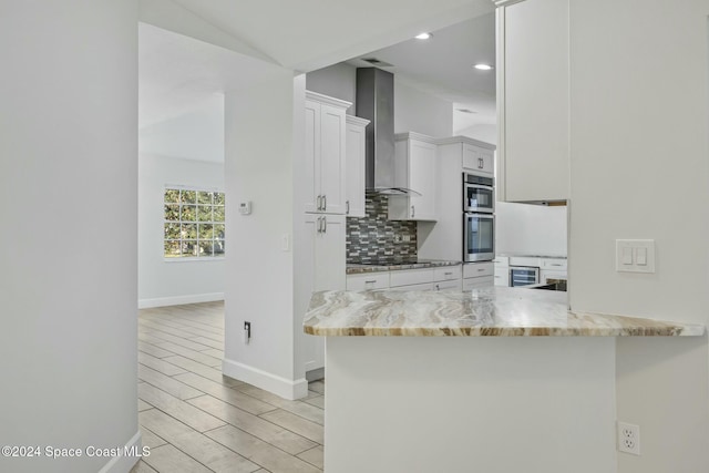 kitchen with kitchen peninsula, light stone counters, white cabinetry, and wall chimney range hood