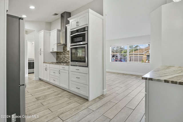 kitchen with light hardwood / wood-style flooring, wall chimney exhaust hood, appliances with stainless steel finishes, light stone counters, and white cabinetry