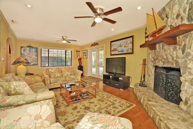 living room with ceiling fan, wood-type flooring, crown molding, a stone fireplace, and french doors