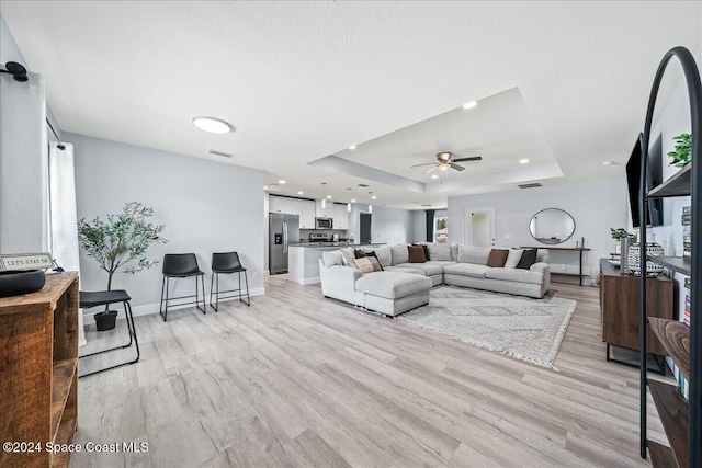 living room with a raised ceiling, ceiling fan, a textured ceiling, and light wood-type flooring