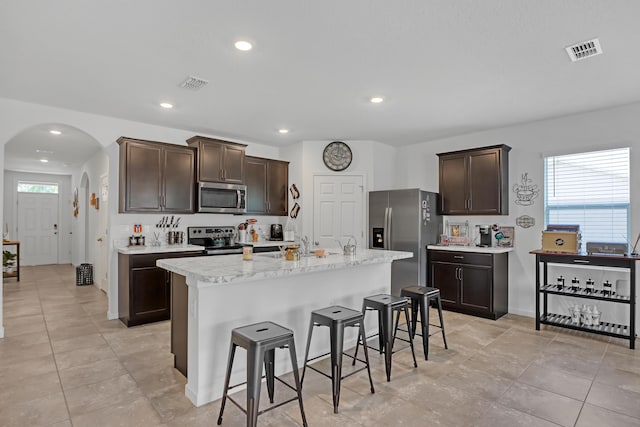 kitchen with a breakfast bar, a center island with sink, light tile patterned floors, dark brown cabinetry, and stainless steel appliances