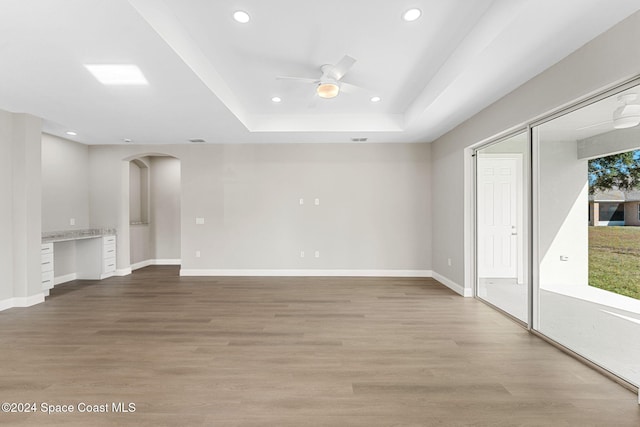 unfurnished living room with ceiling fan, a raised ceiling, built in desk, and light hardwood / wood-style flooring