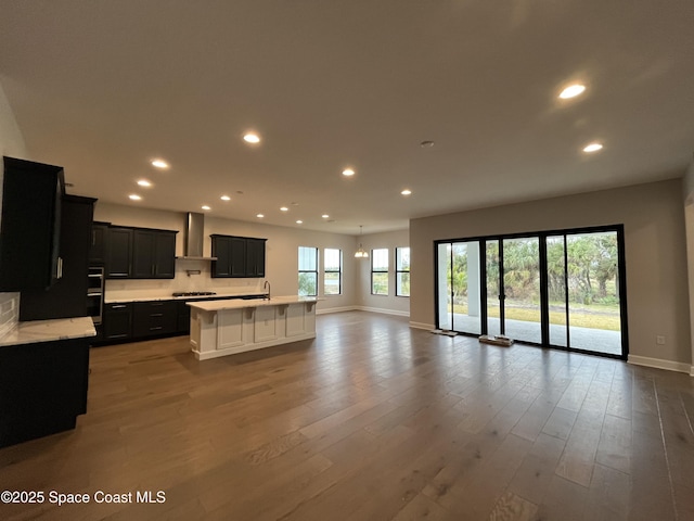 kitchen featuring light hardwood / wood-style floors, a center island with sink, gas stovetop, wall chimney range hood, and double oven