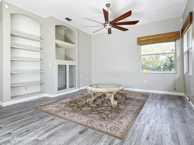 living area featuring built in shelves, ceiling fan, wood-type flooring, and a textured ceiling