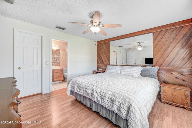 bedroom featuring wooden walls, ceiling fan, light wood-type flooring, a textured ceiling, and connected bathroom