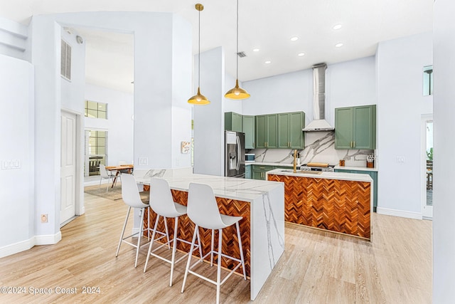 kitchen featuring stainless steel refrigerator with ice dispenser, wall chimney exhaust hood, pendant lighting, a high ceiling, and green cabinets