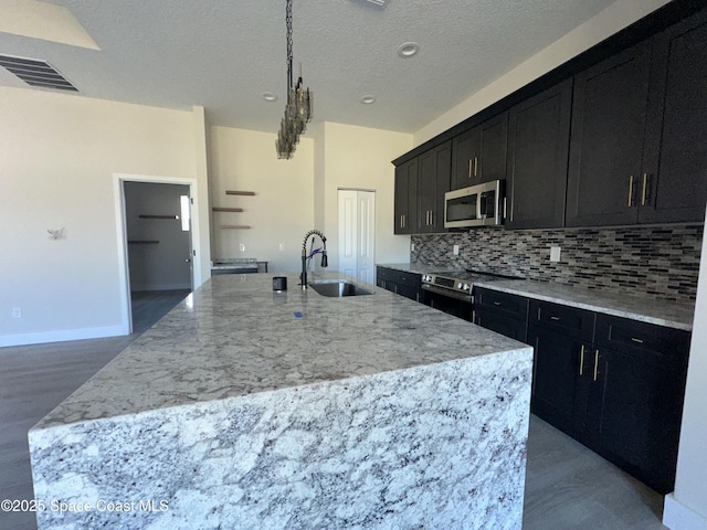 kitchen with appliances with stainless steel finishes, tasteful backsplash, sink, light stone countertops, and a center island with sink